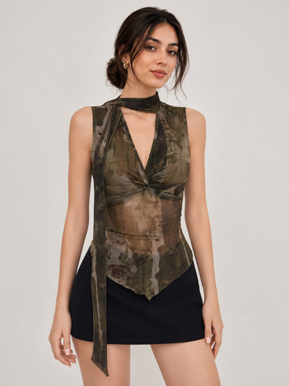 A woman modeling a sleeveless high-neck top with a tie-front design and a distressed camouflage print in olive green, brown, and black, paired with a black mini skirt against a light gray studio background.