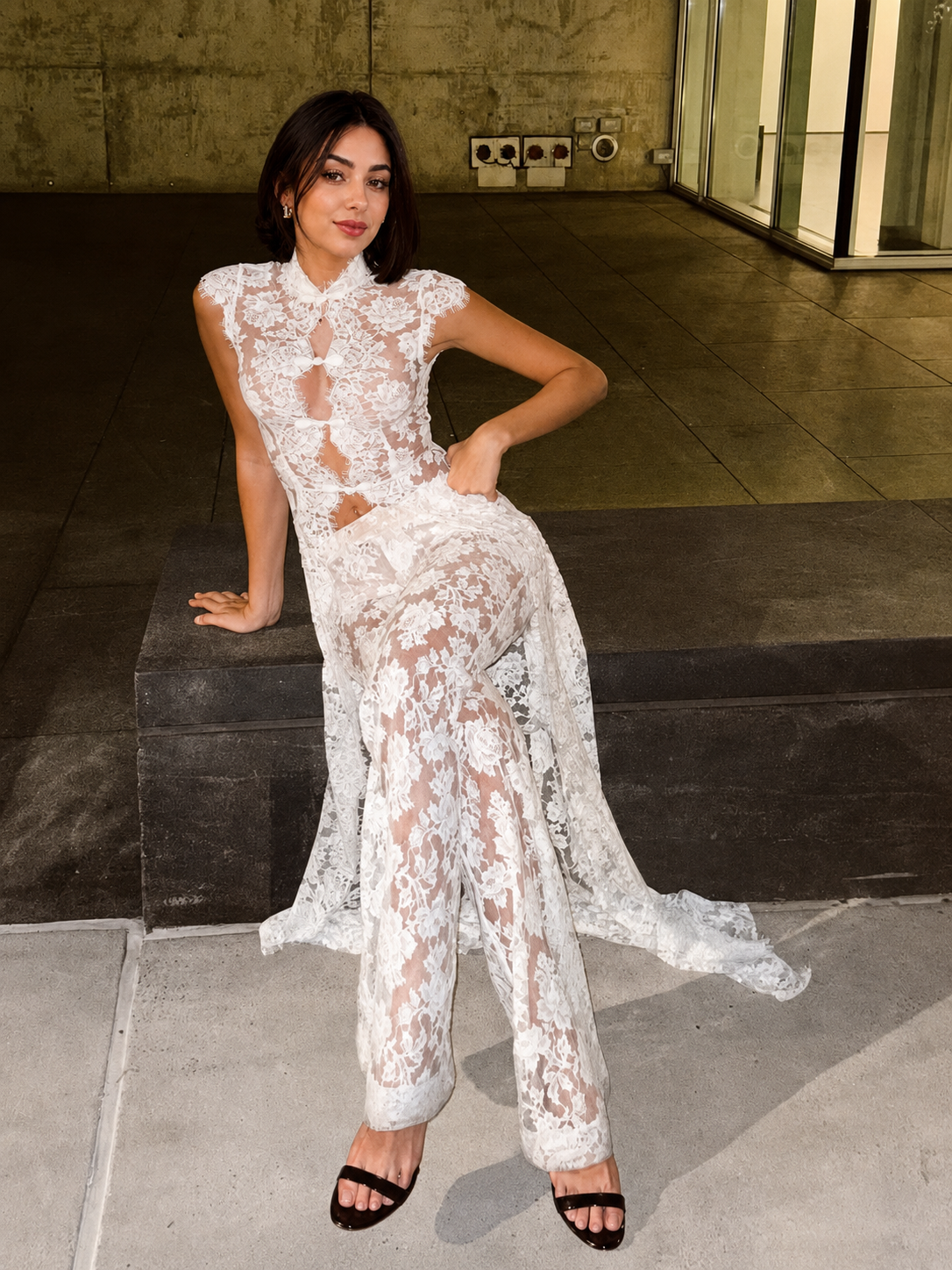 A woman wearing a sheer white lace maxi dress with a high collar and cutout details, posing on dark stone steps in an outdoor urban setting.