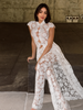 A woman wearing a sheer white floral lace jumpsuit with cap sleeves and a high collar, posing on stone steps against a textured concrete wall.