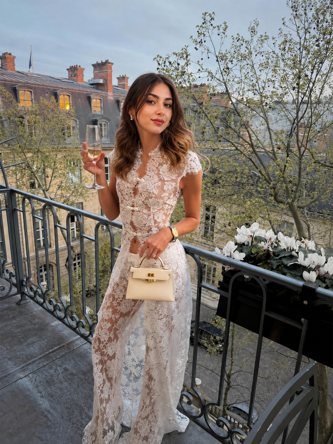 A woman stands on an outdoor balcony wearing a sheer white lace dress and holding a small beige handbag, with a cityscape and trees in the background.