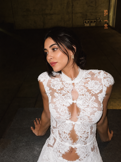 A woman wearing a white lace dress with sheer floral patterns and cutout details, photographed in a dim indoor setting.