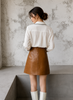 Woman wearing a white blouse and brown leather skirt against a textured wall.