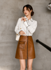 Woman wearing a white blouse with black trim and a brown leather skirt against a gray wall.