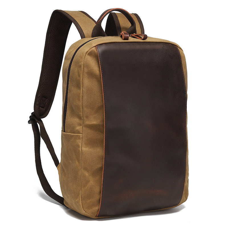 Canvas laptop backpack carried by hand, showing tan waxed canvas, dark brown contrast section, top handle, and shoulder straps.