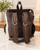 A dark brown faux-leather backpack with wide brown webbed shoulder straps, silver D-rings, and a cowhide-textured side panel, shown on a marble countertop.