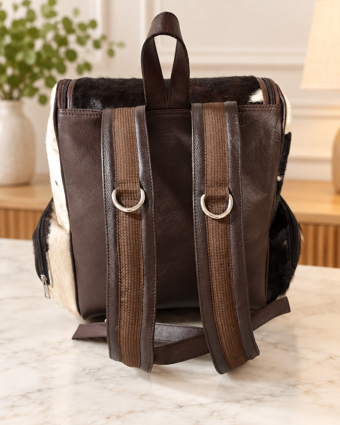 A dark brown faux-leather backpack with wide brown webbed shoulder straps, silver D-rings, and a cowhide-textured side panel, shown on a marble countertop.