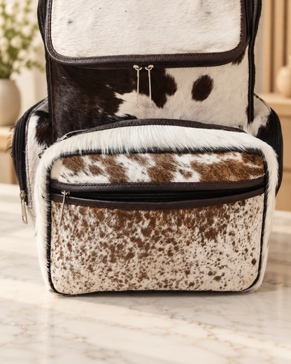 Cowhide mommy backpack with brown leather flap opened above natural hair-on hide and visible zippered storage compartments.