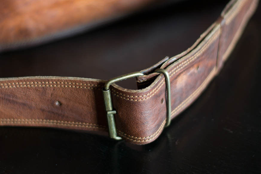 A rugged leather men’s messenger bag in distressed brown, paired with a journal and watch on a rustic wooden surface.