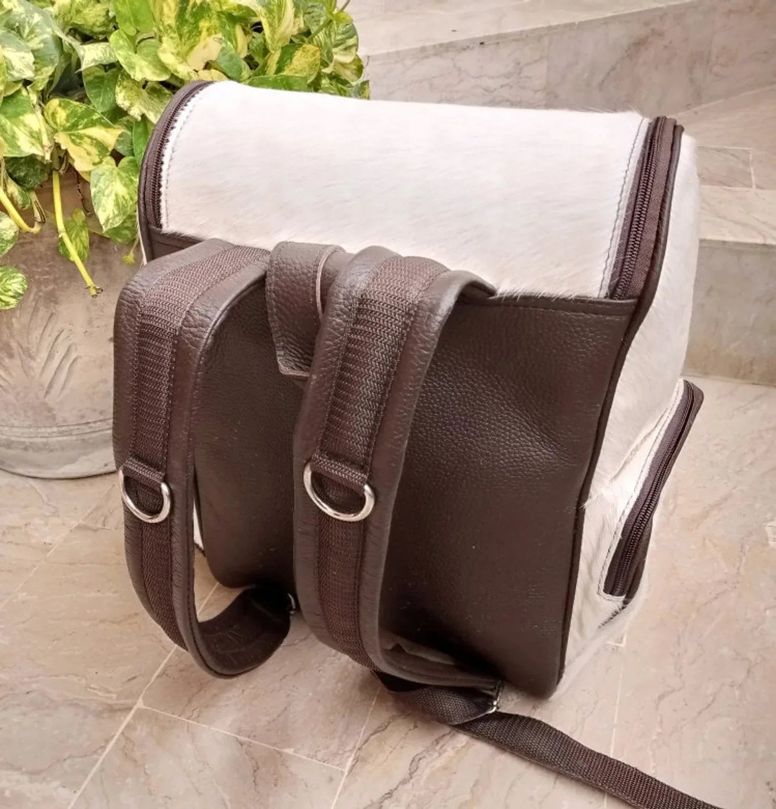 Brown cowhide backpack with white natural markings, hair-on hide exterior, front zipper pockets, and dark leather backing.