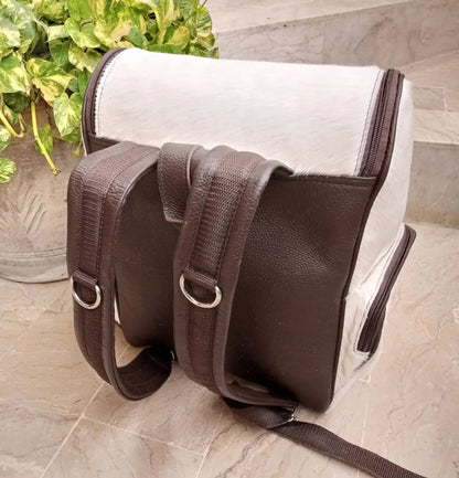 Brown cowhide backpack with white natural markings, hair-on hide exterior, front zipper pockets, and dark leather backing.