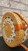 Round rattan bag with intricate woven design, resting on a wooden table, sunlight casting soft shadows.