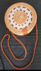 Handwoven rattan purse with a circular design, adorned with a leather strap, resting on a light, textured surface.