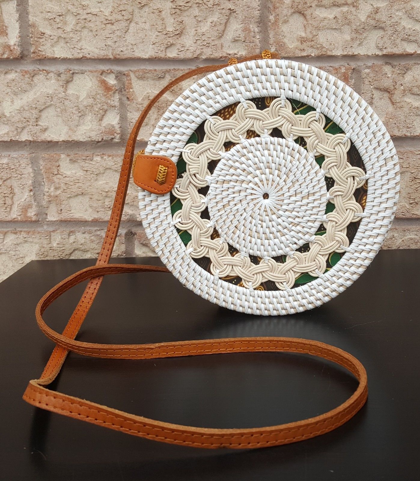 Round rattan bag with intricate woven design, leather strap, and clasp, placed on a rustic wooden surface