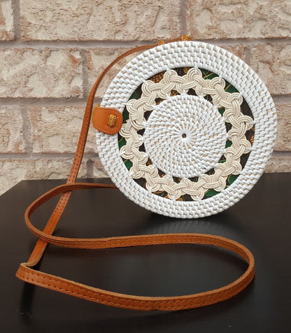 Round rattan bag with intricate woven design, leather strap, and clasp, placed on a rustic wooden surface